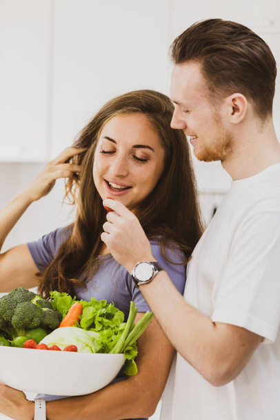 Optimistic couple with bowl full of assorted raw healthy vegetables at table in light kitchen together with modern appliances in apartment on vita - Photo, Image