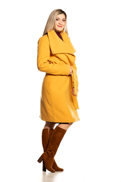 Молодая красивая блондинка в желтом пальто и кожаных сапогах на белом фоне - Фото, изображение