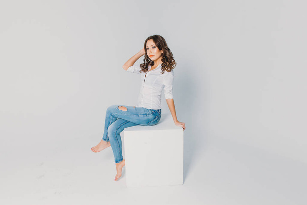 a girl in a white shirt and jeans sits on a white cube on a white background - 写真・画像