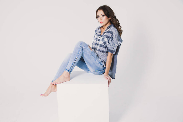 a girl in a plaid shirt and jeans sits on a white cube on a white background - Zdjęcie, obraz