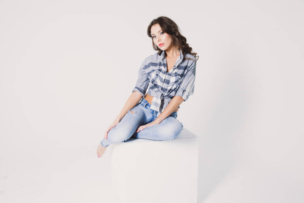 a girl in a plaid shirt and jeans sits on a white cube on a white background - Fotografie, Obrázek