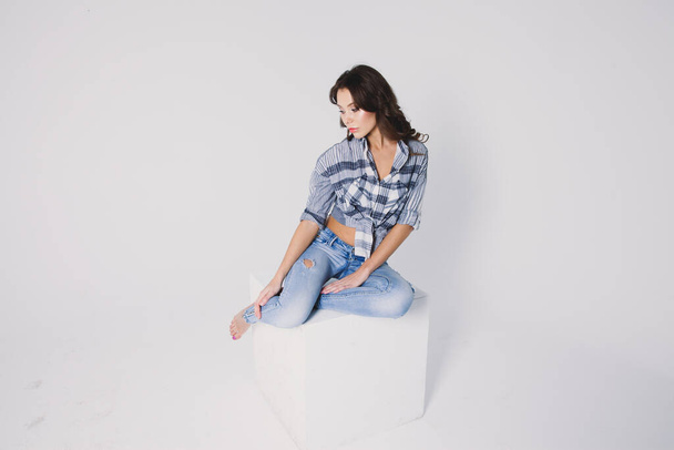 a girl in a plaid shirt and jeans sits on a white cube on a white background - Φωτογραφία, εικόνα