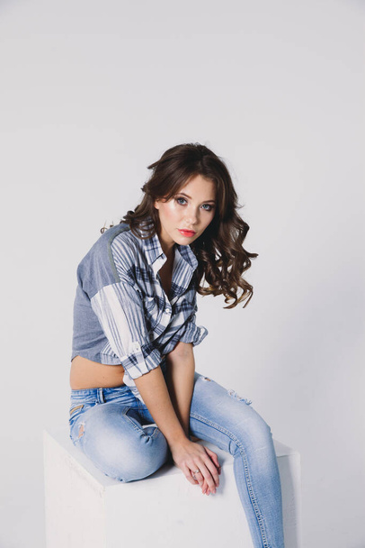 a girl in a plaid shirt and jeans sits on a white cube on a white background - Zdjęcie, obraz