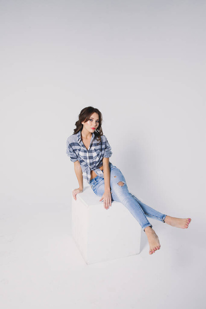 a girl in a plaid shirt and jeans sits on a white cube on a white background - Fotoğraf, Görsel