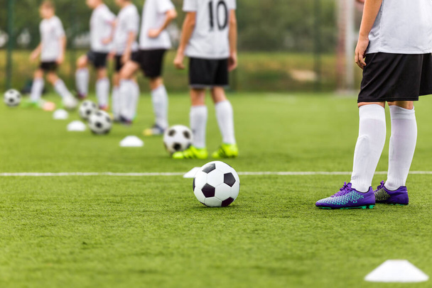 サッカー選手たちはトレーニングユニットのサッカーボールと並んで立っている。子供のためのサッカースクール。草のピッチでサッカーを練習している若い男の子。白と黒のサッカーキットの子供たち - 写真・画像