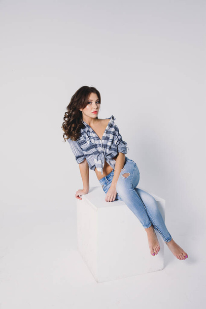 a girl in a plaid shirt and jeans sits on a white cube on a white background - Фото, зображення
