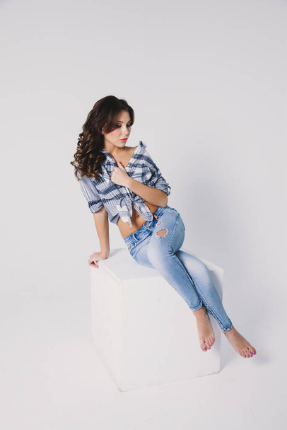 a girl in a plaid shirt and jeans sits on a white cube on a white background - Foto, Imagem
