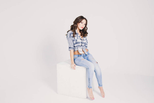 a girl in a plaid shirt and jeans sits on a white cube on a white background - Φωτογραφία, εικόνα