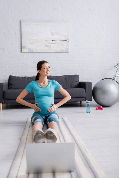 счастливая беременная женщина в спортивной одежде упражнения возле ноутбука на фитнес-коврик - Фото, изображение