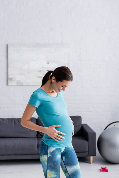cheerful pregnant sportswoman touching belly in living room  - Photo, Image
