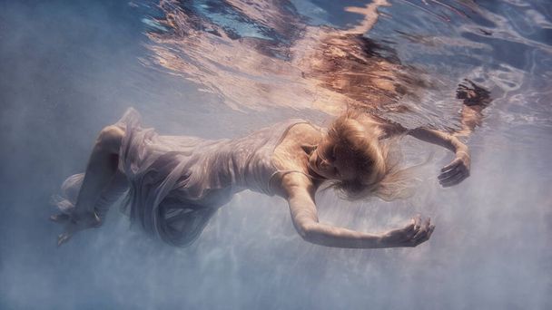     A woman with blond hair in a white dress swims underwater as if flying in zero gravity                           - Photo, image
