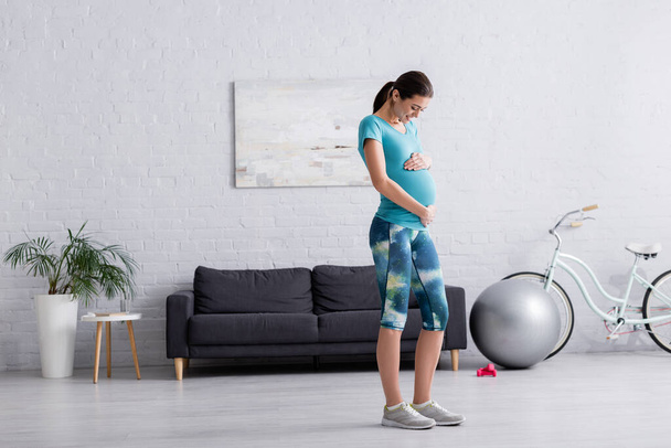 full length of happy pregnant woman touching belly in living room  - Foto, Bild