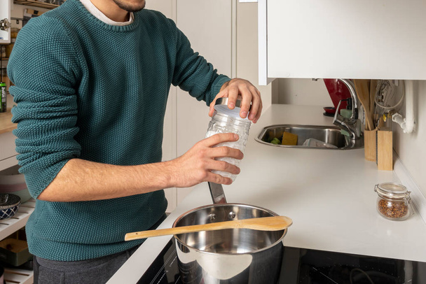 Руки молодого чоловіка відкручують банку морської солі, щоб приправити їжу, яку він збирається приготувати на скляній керамічній плиті
 - Фото, зображення