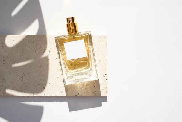Transparante fles parfum met wit etiket op stenen plaat op een witte achtergrond. Parfumpresentatie met daglicht. Trending concept in natuurlijke materialen met palmblad schaduwen. - Foto, afbeelding