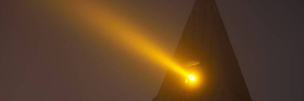 Una antigua iglesia del faro histórico por la noche. Letonia. Rayo de luz dorada contra cielo claro del crepúsculo. Paisaje urbano oscuro. Lugares de interés, turismo, destinos turísticos. Símbolo de esperanza y paz - Foto, imagen