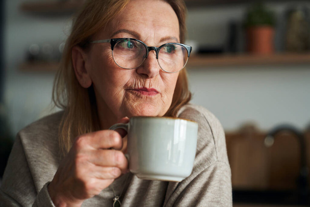 Close up of thoughtful caucasian senior woman sitting in the kitchen, drinking coffee and looking away - 写真・画像