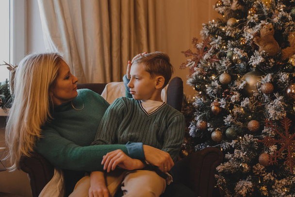 Piękna kobieta matka i syn siedzą na krześle przy choince w noworocznym domu - Zdjęcie, obraz