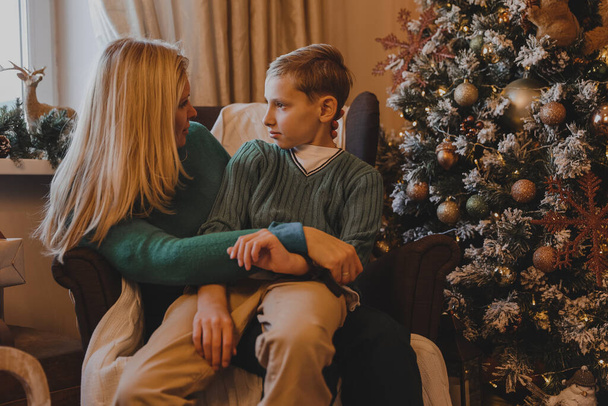 Όμορφη γυναίκα μητέρα και γιος κάθονται σε μια καρέκλα δίπλα στο χριστουγεννιάτικο δέντρο στο στολισμένο σπίτι της Πρωτοχρονιάς. - Φωτογραφία, εικόνα