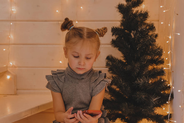 Cute girl speaks on a smartphone in a Christmas atmosphere, a New Year's decorated house, Children and modern technology, communication concept. - Фото, изображение