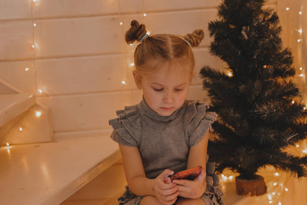 Cute girl speaks on a smartphone in a Christmas atmosphere, a New Year's decorated house, Children and modern technology, communication concept. - Foto, Imagen