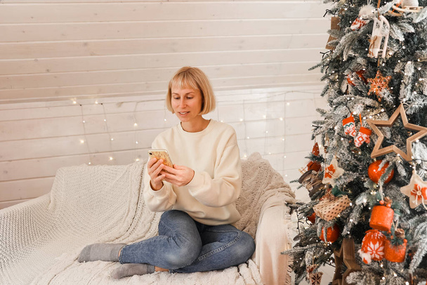 Şirin bir kadın Noel atmosferinde akıllı bir telefonla konuşuyor. Yeni yıl dekore edilmiş bir ev, modern teknoloji, iletişim konsepti.. - Fotoğraf, Görsel