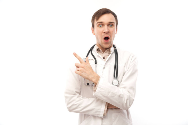 Handsome doctor in white medical coat with stethoscope points finger at empty space isolated on white background with copy space. Demonstrates product, excitedly shows something - Zdjęcie, obraz