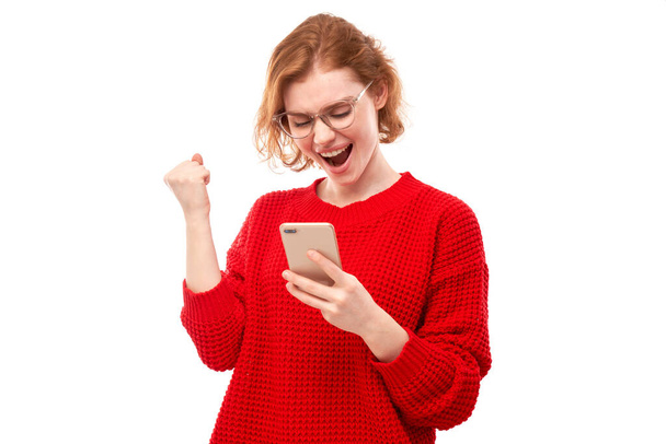 lucky smiling redhead girl holding smartphone in red clothes with glasses isolated in studio received joyful message, celebrating victory - Φωτογραφία, εικόνα