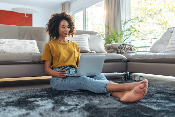 Снимок зрелой расслабляющей женщины, платящей что-то онлайн с помощью своей кредитной карты с ноутбуком, сидя на полу в гостиной дома. - Фото, изображение
