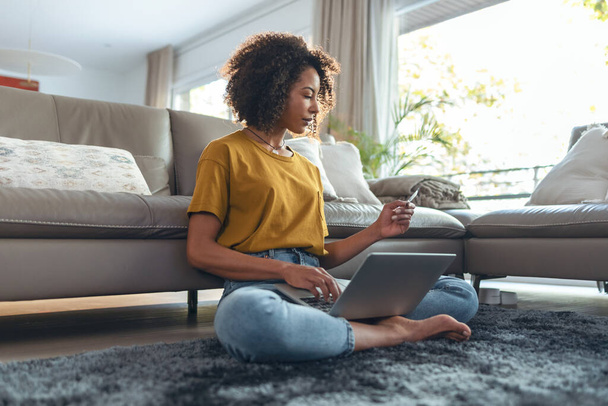 Shot of mature relaxing woman paying something online with her credit card with laptop while sitting on the floor in living room at home. - Фото, изображение