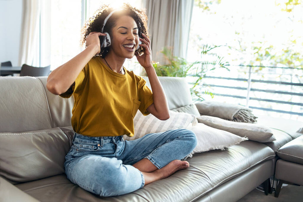 Shot of motivated mature woman listening to music with headphones on her smartphone while dancing and singing on sofa at home. - Foto, afbeelding