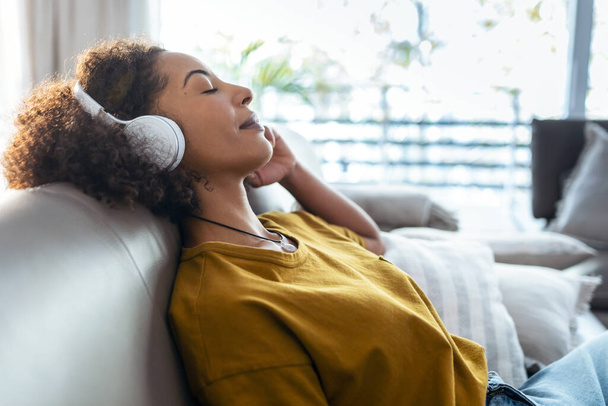 Shot of beautiful mature woman listening to music with headphones on her smartphone while relaxing sitting on sofa at home. - Fotó, kép