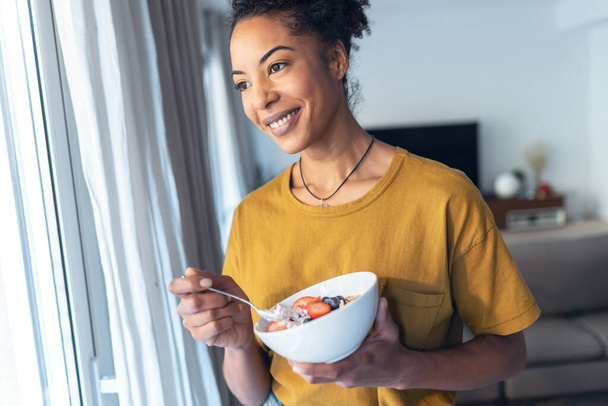 Shot of beautiful mature woman eating cereals and fruits while standing next to the window at home. - Foto, afbeelding
