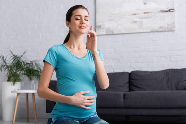 mulher grávida tatuada com olhos fechados meditando na sala de estar  - Foto, Imagem