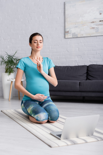joven embarazada con los ojos cerrados meditando en la sala de estar  - Foto, Imagen