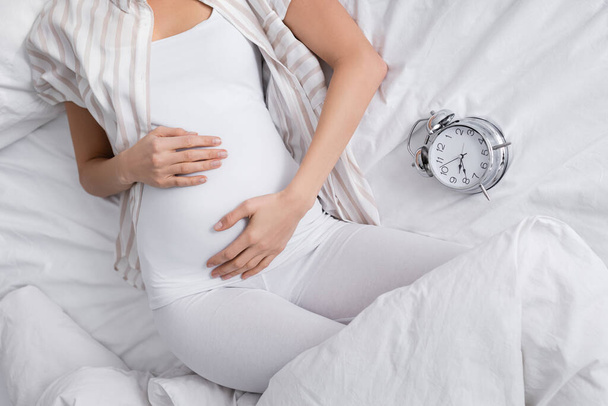 cropped view of pregnant woman near alarm clock on bed - Fotoğraf, Görsel