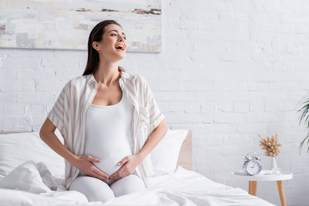 onnellinen raskaana oleva nainen nauraa ja koskettaa vatsa makuuhuoneessa - Valokuva, kuva