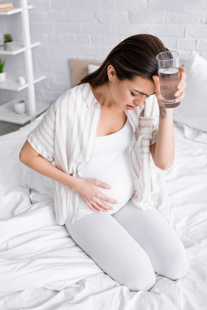 jonge zwangere vrouw die een glas water vasthoudt terwijl ze buikpijn voelt  - Foto, afbeelding