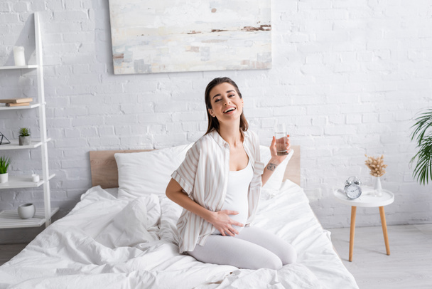 cheerful pregnant woman holding glass of water in bedroom - Фото, изображение