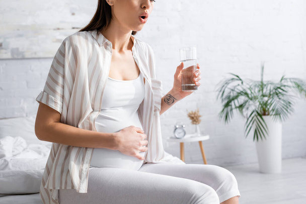 vista recortada de la mujer embarazada sosteniendo un vaso de agua mientras siente dolor abdominal  - Foto, Imagen