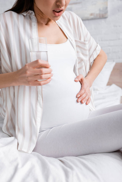 bijgesneden zicht op zwangere vrouw die kramp voelt terwijl ze glas water vasthoudt - Foto, afbeelding