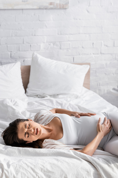 mujer embarazada joven con los ojos cerrados que sufren de calambres mientras está acostada en la cama - Foto, Imagen