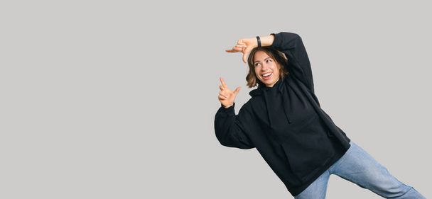 Young cheerful woman in black hoodie pointing over grey background at copyspace. - Фото, зображення