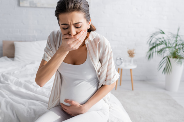 νεαρή έγκυος γυναίκα αισθάνεται ναυτία και καλύπτει το στόμα στο υπνοδωμάτιο  - Φωτογραφία, εικόνα