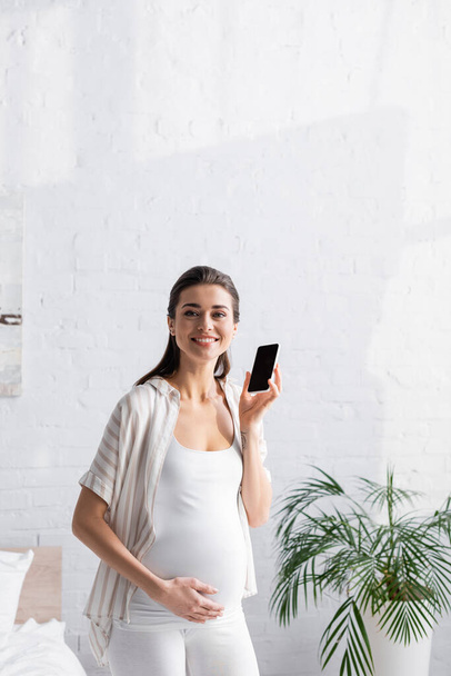 donna incinta sorridente che tiene smartphone in camera da letto  - Foto, immagini