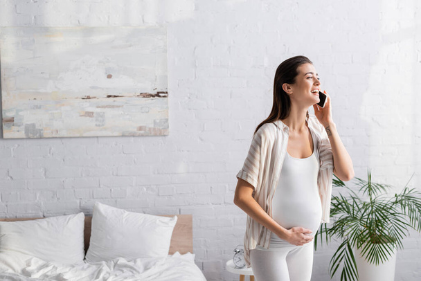 positive pregnant woman talking on smartphone in bedroom  - Photo, Image