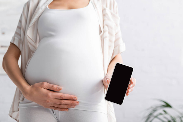 обрізаний вид вагітної молодої жінки, що тримає смартфон з порожнім екраном
 - Фото, зображення