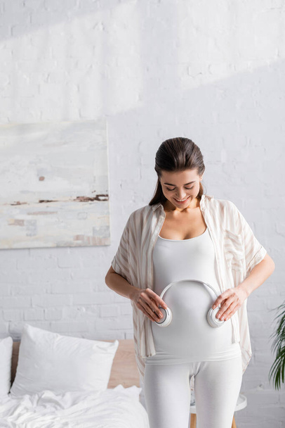happy pregnant woman holding wireless headphones near belly - Photo, image