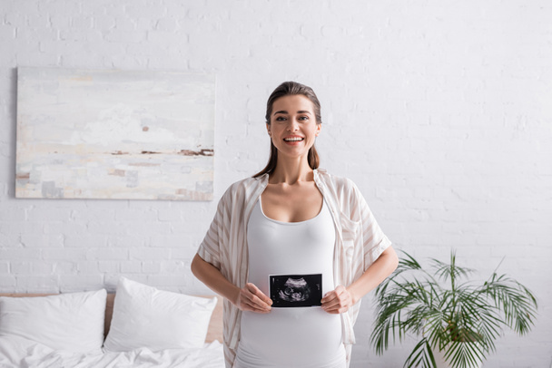 mosolygós és terhes nő kezében ultrahang vizsgálat a hálószobában  - Fotó, kép