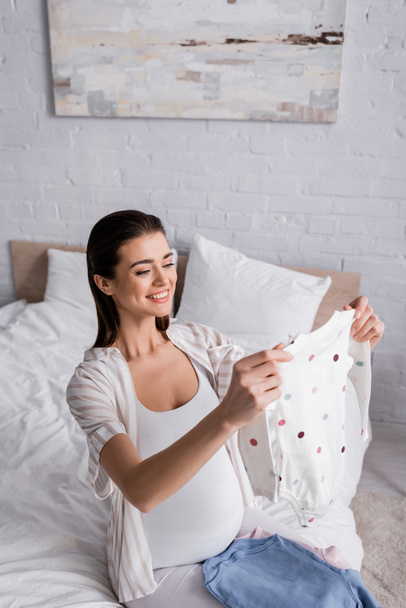 smiling pregnant woman holding baby romper in bedroom  - 写真・画像
