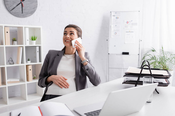 vrolijk zwanger zakenvrouw praten op smartphone in de buurt van laptop op het bureau - Foto, afbeelding